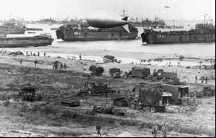 Buques de desembarco de tanques (LST) y otros buques de la Armada de EE. UU. descargando durante la marea baja en Normandía, poco después de la invasión de junio de 1944. El USS LST-55 está en el centro, detrás del globo de barrera más cercano. El USS LST-61 está a la derecha. Crédito: Steck, Cuerpo de Señales del Ejército de EE. UU., dominio público, vía Wikimedia Commons.